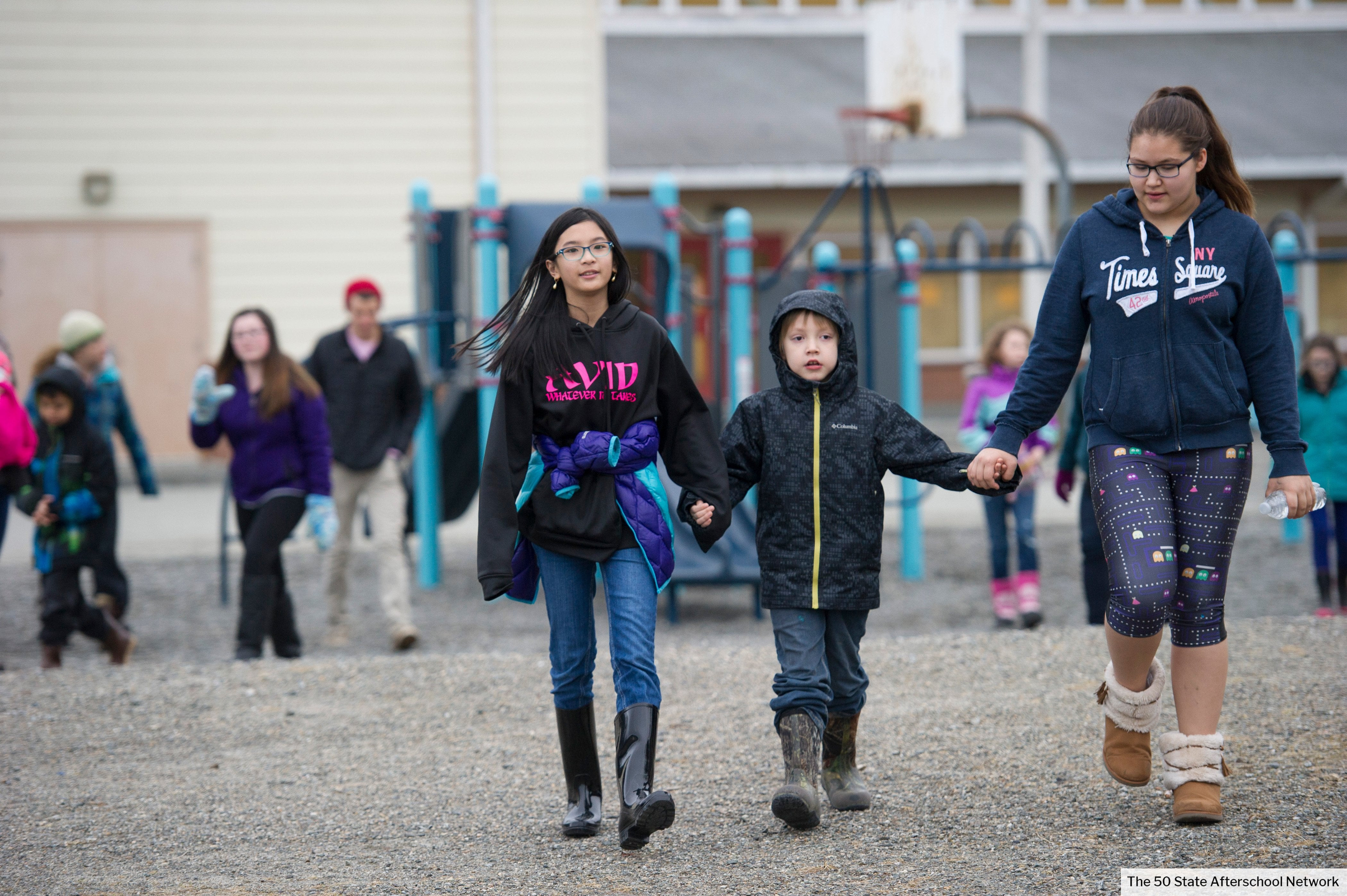 One young female-presenting Asian student and one young female-presenting Latinx student are holding hands with a young white male-presenting student as they are walking in a school playground.