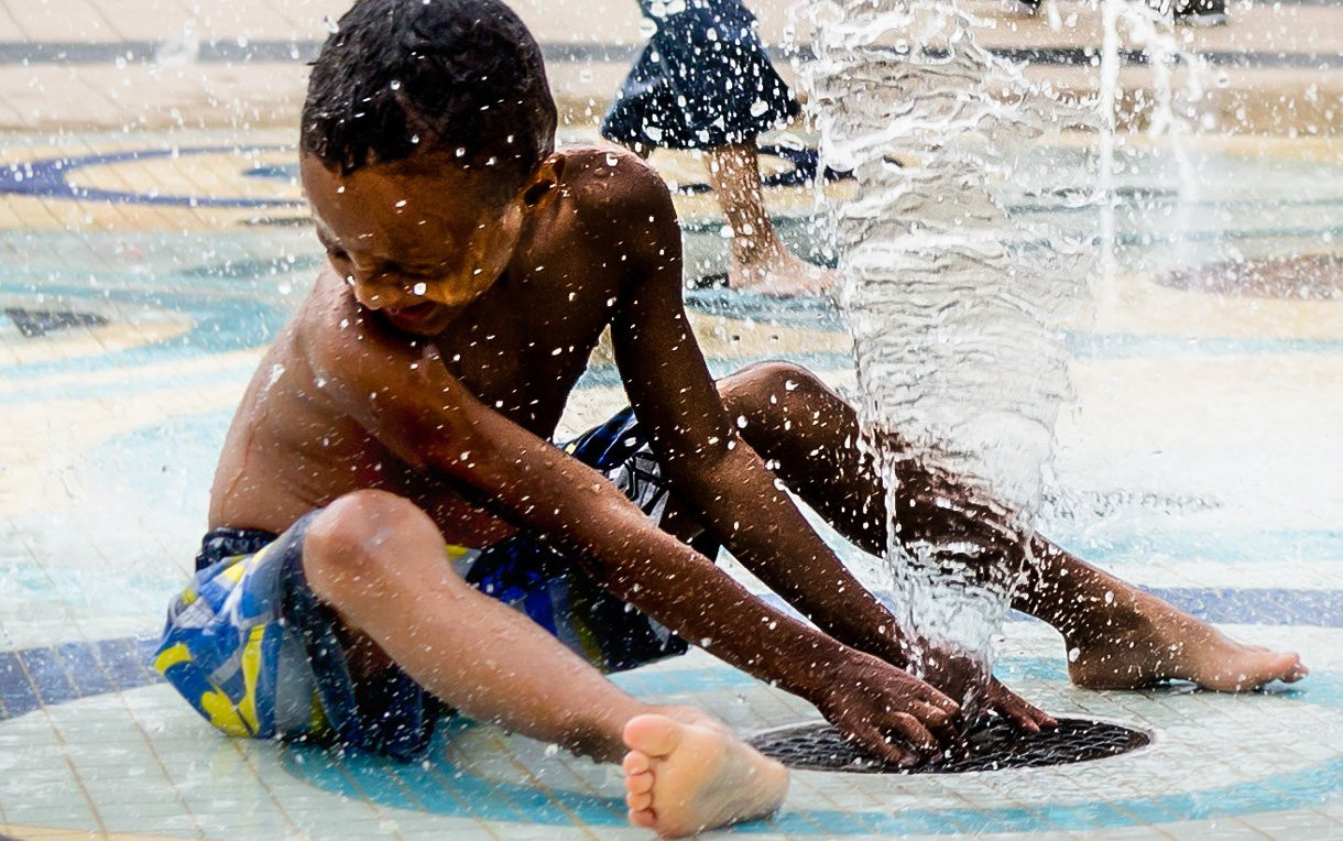 A kid sits on the ground and plays with a stream of water at a splash park.