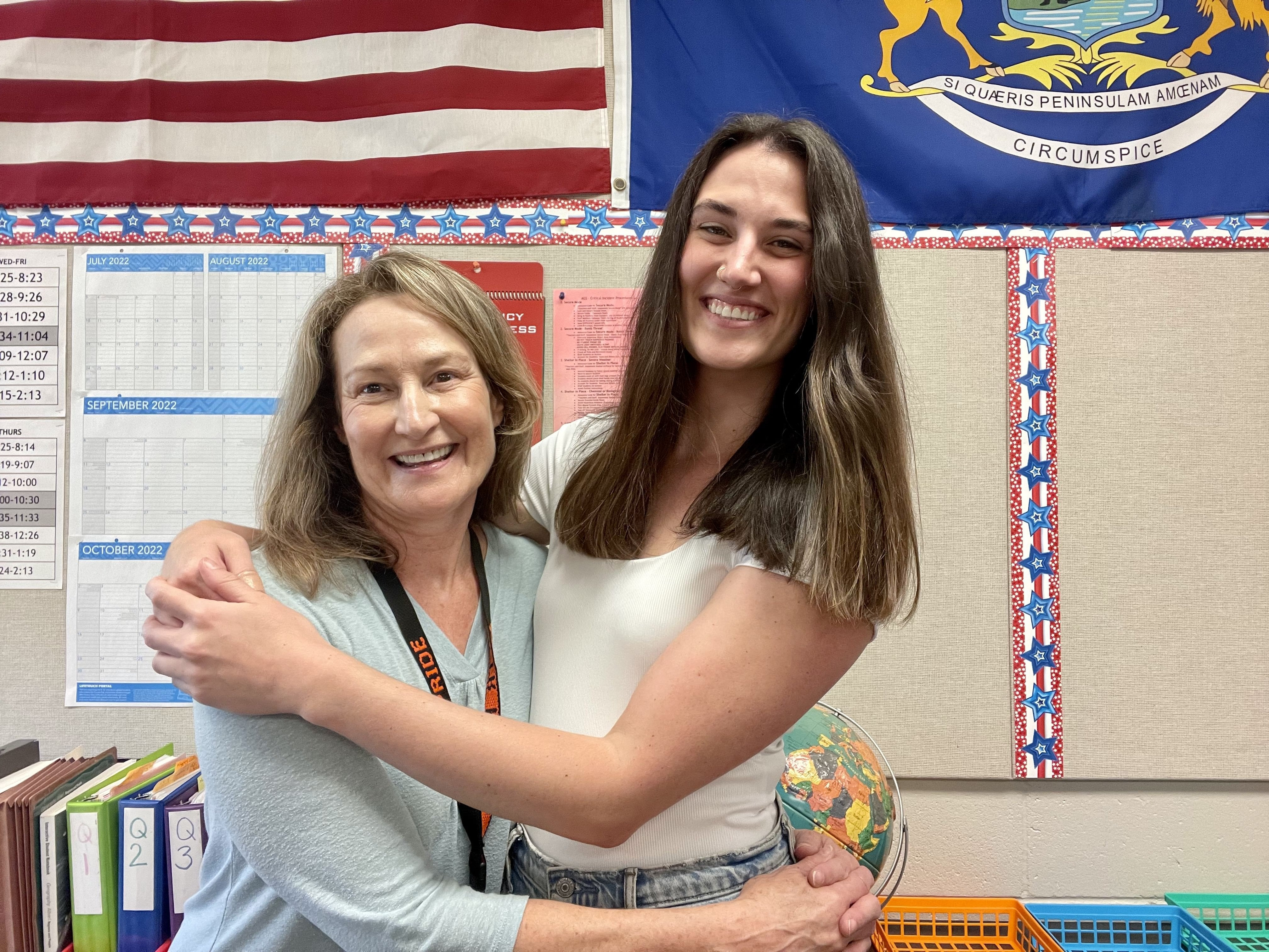Two white women hugging each other sideways in a school classroom.