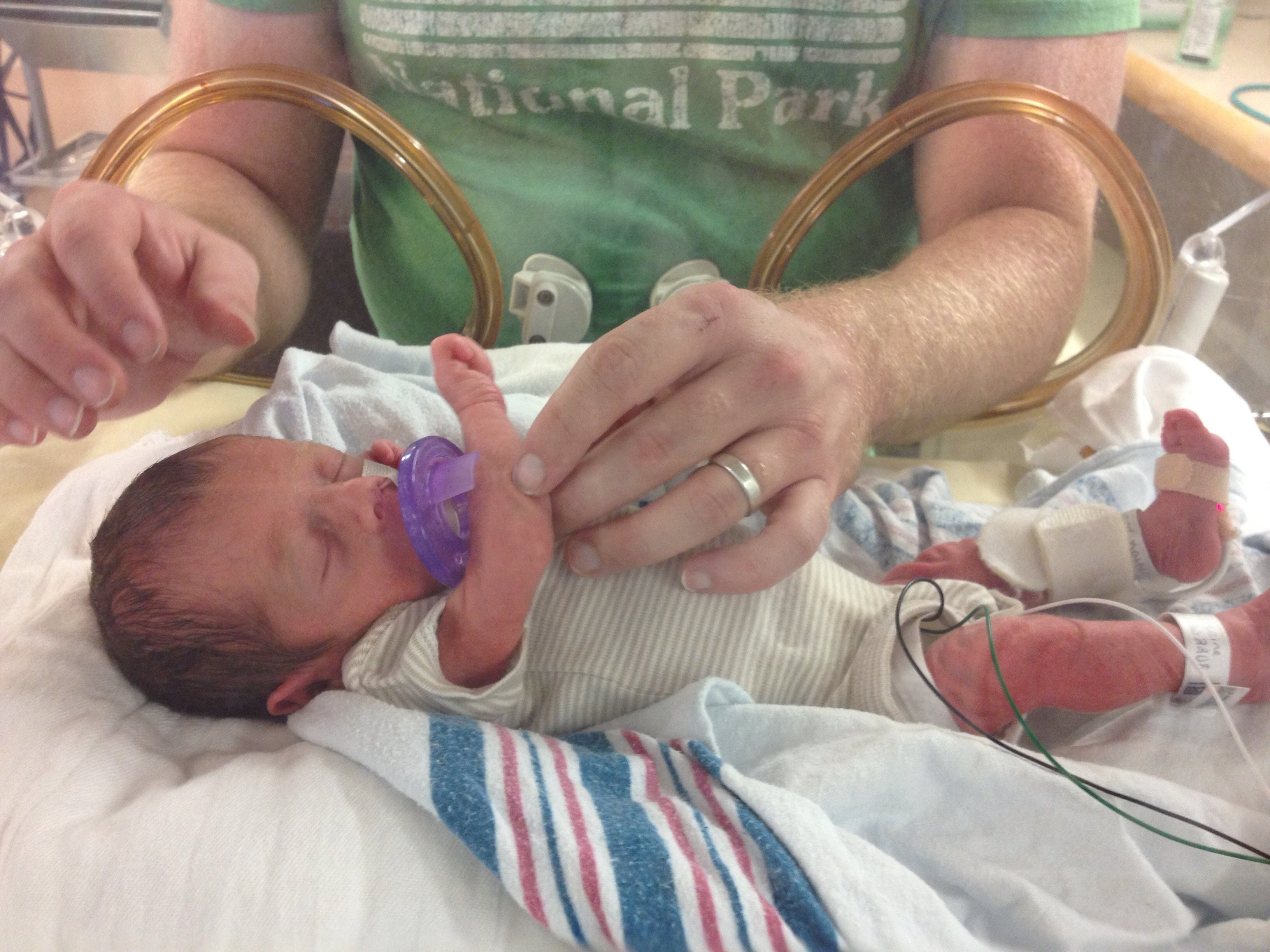 An adult male wearing a green shirt is tending a premature baby in the NICU.
