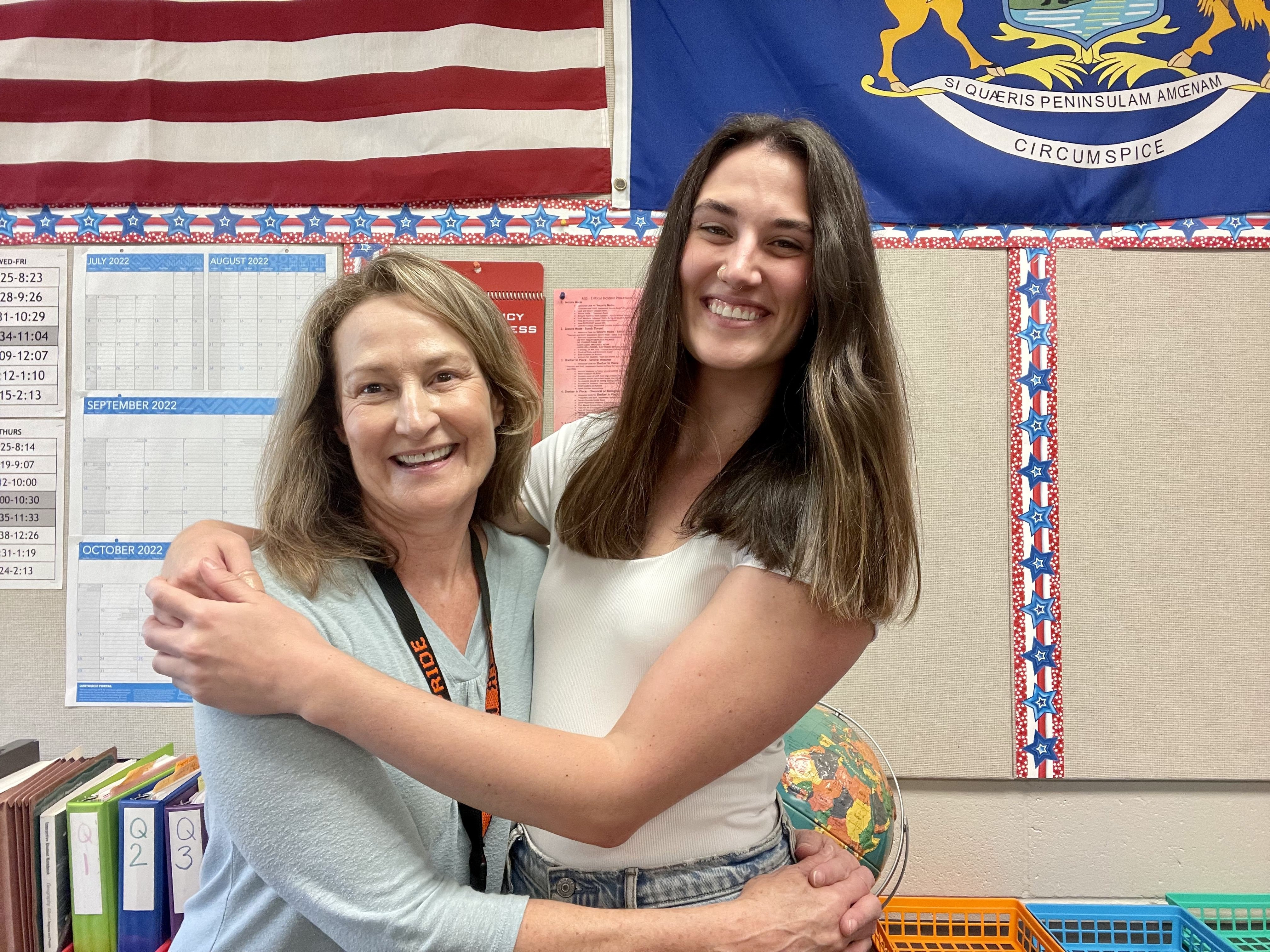 Two white women hugging each other sideways in a school classroom.