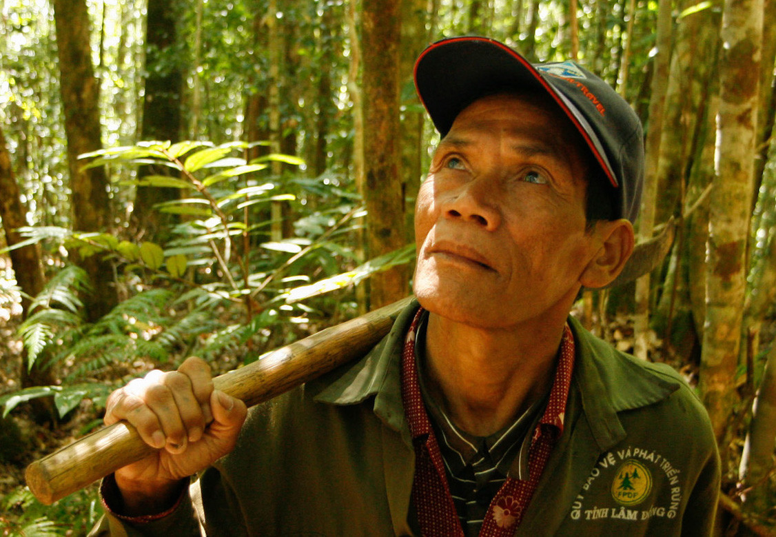 A man standing in a forest with a hat on, holding a stick over his back.