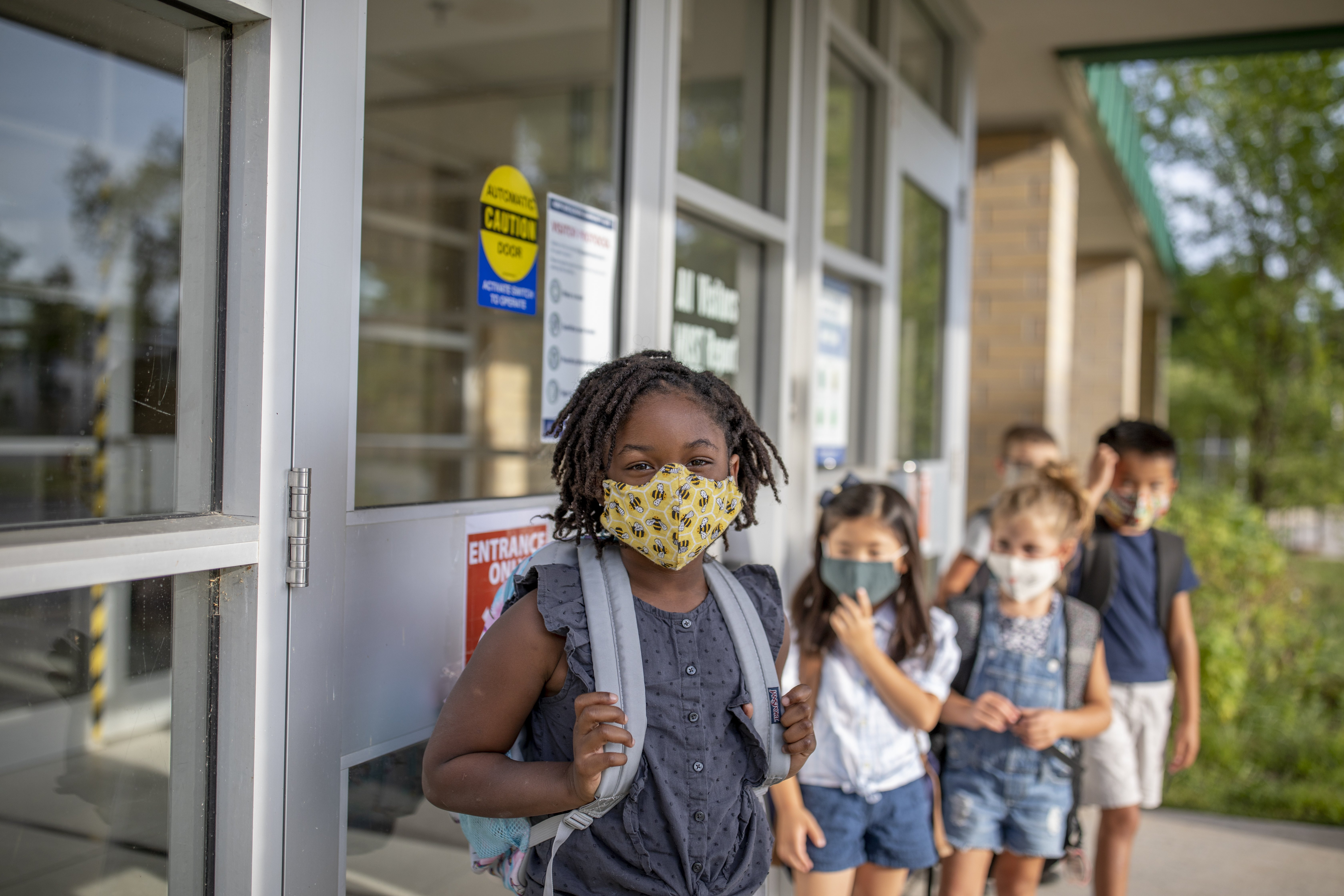 A young Black girl with a yellow mask with bees wearing a gray blouse is smiling at the camera. Her classmates from diverse ethnic backgrounds are standing in the background with their masks on.