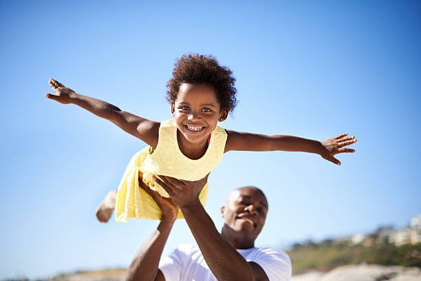 A Black father holding his Black daughter up in a flying position.