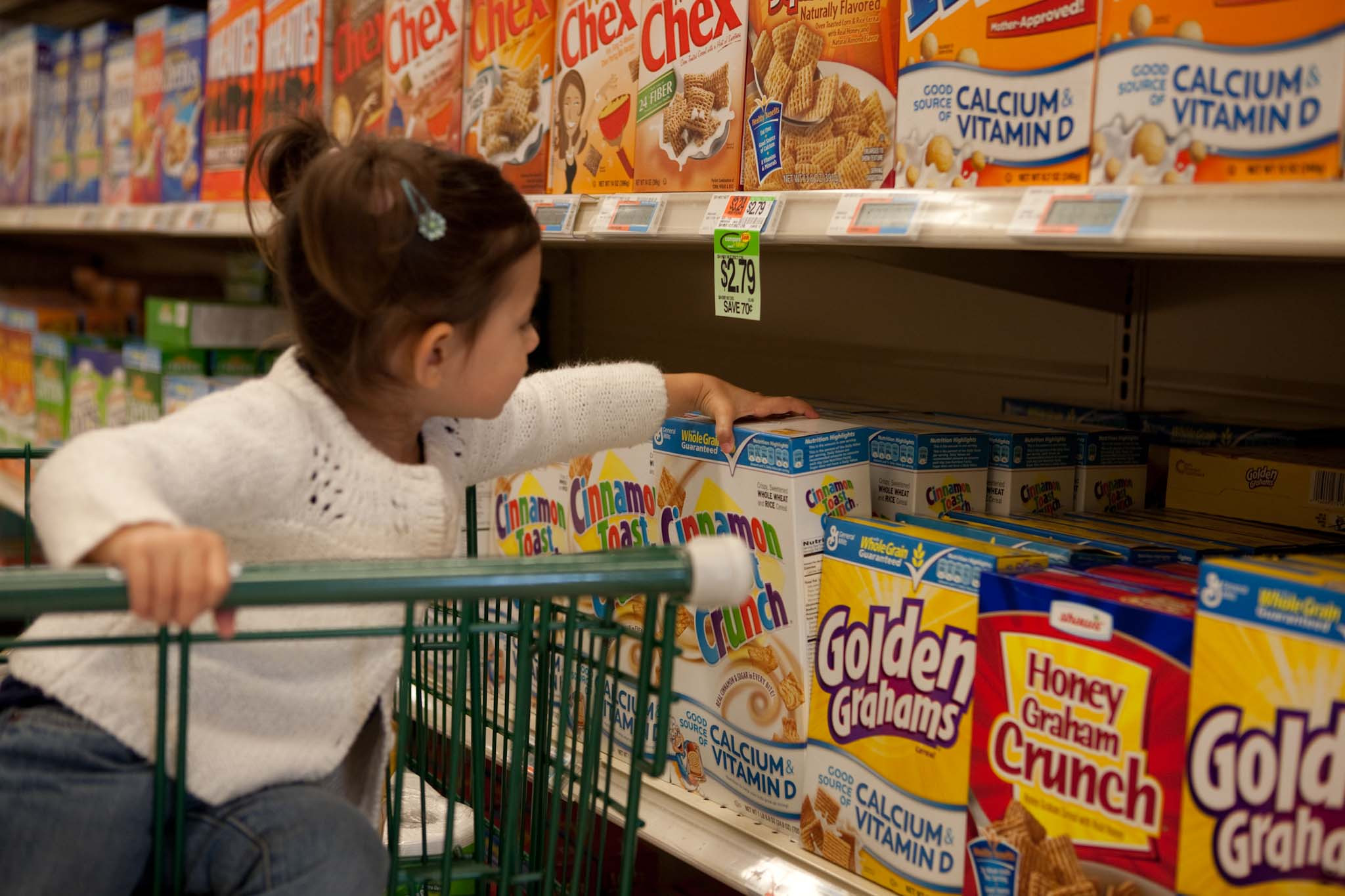 Toddler with hair clips wearing a white sweater and jeans grabbing a box of Cinnamon Toast Crunch off a supermarket shelf.