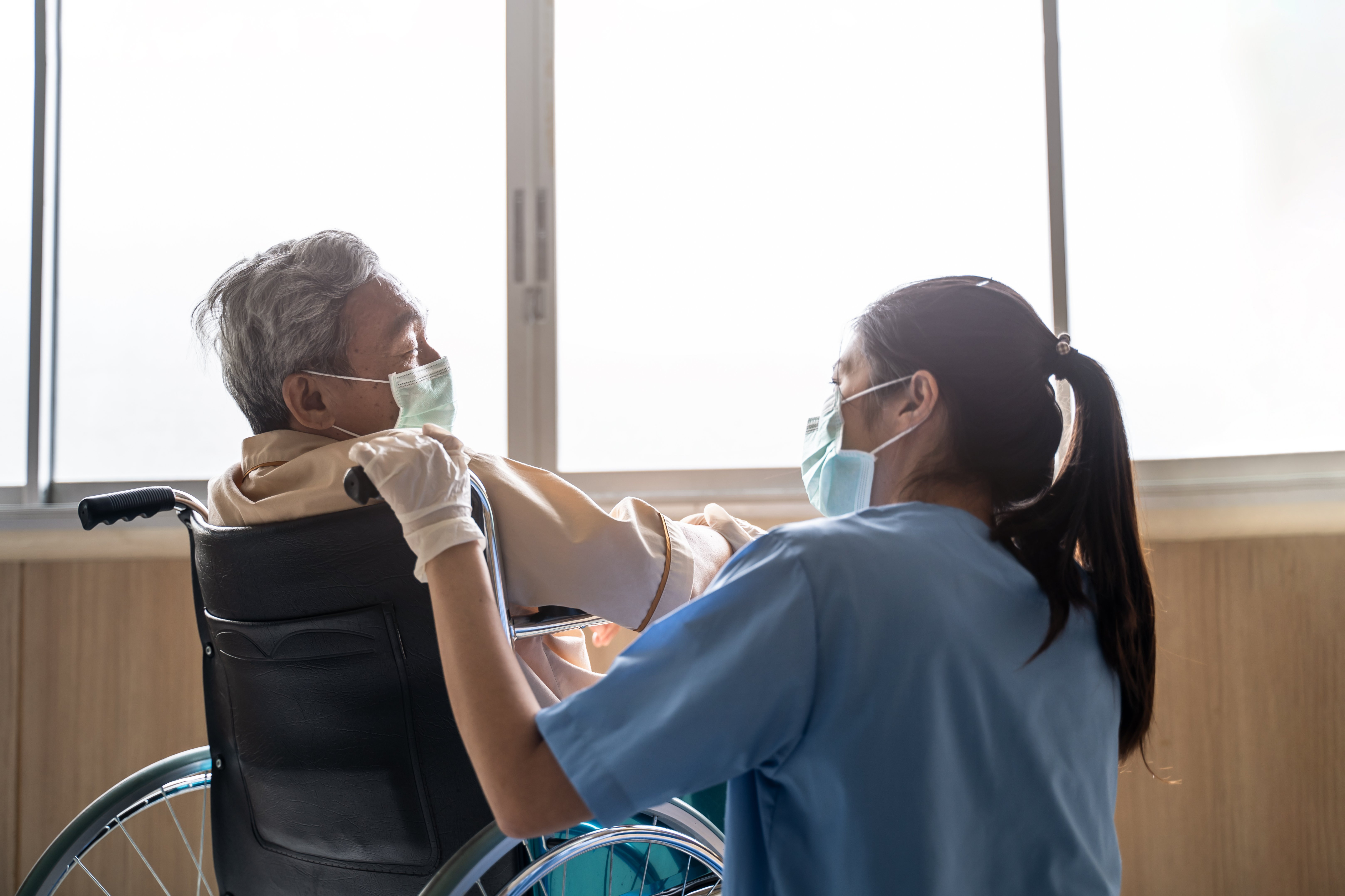 An Asian female nurse in scrubs and a mask is looking endearingly at an elderly male patient who is seated in a wheelchair.